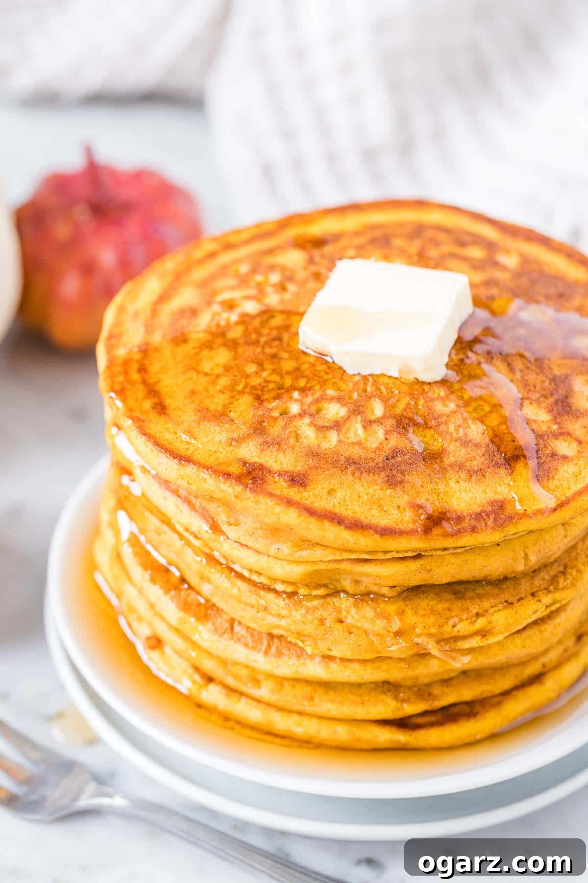 A tall stack of golden Pumpkin Pancakes, generously topped with a pat of melting butter and drizzled with rich maple syrup, ready to be enjoyed.