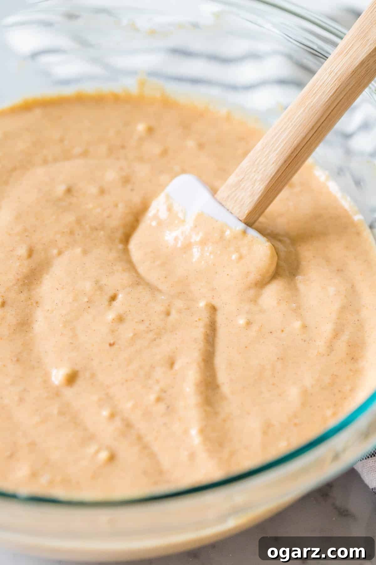 A glass bowl containing a smooth, creamy pumpkin pancake batter, with a spatula resting inside, ready to be cooked.