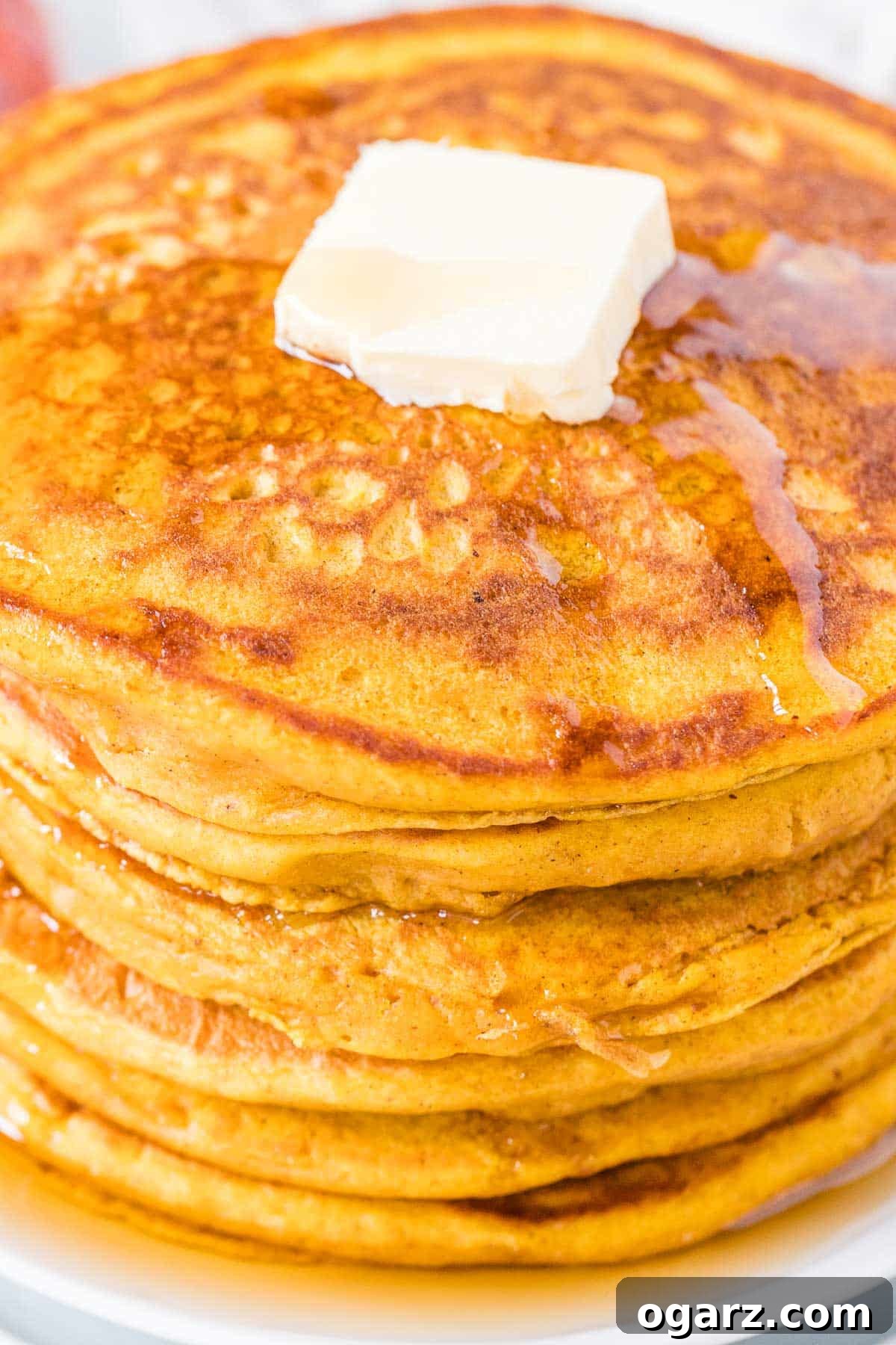 A close-up view of freshly cooked pumpkin pancakes, stacked high on a white plate, showcasing their fluffy texture and golden-brown edges.