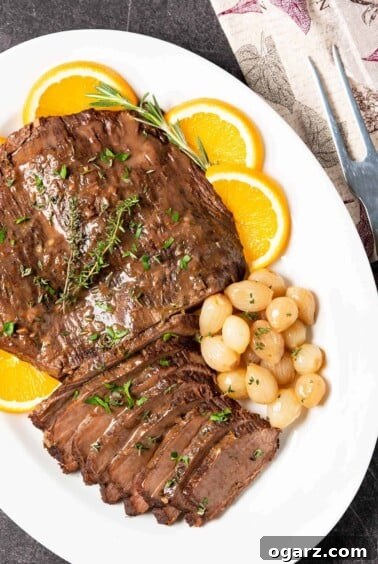 A serving platter of partially carved beef brisket garnished with oranges, pearl onions, fresh rosemary and thyme.