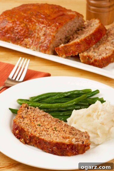 A slice of meatloaf on a dinner plate with mashed potatoes and green beans, a serving platter of meatloaf in the background.