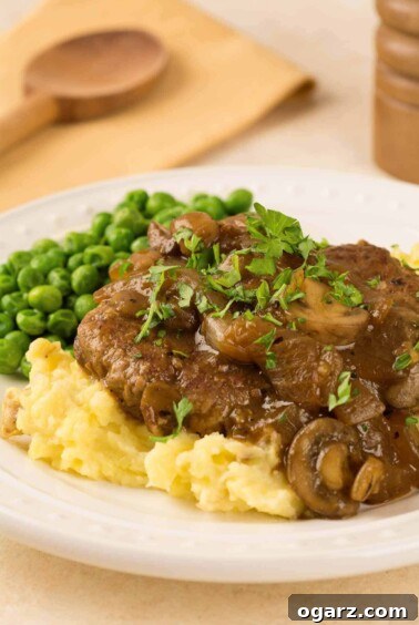 Salisbury steak topped with mushroom and onion gravy, served with mashed potatoes and peas on a white plate.