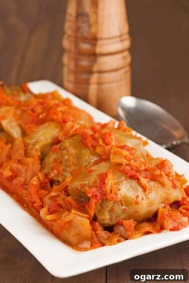 A half dozen stuffed cabbage rolls on a white serving platter, surrounded by chopped cabbage, a serving spoon and pepper mill in the background.