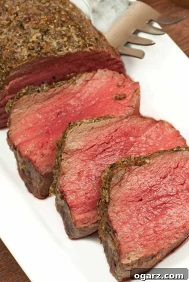 A beautifully roasted beef tenderloin, partially carved, sits on a pristine white plate, showcasing its tender, pink interior and a rich, glossy port wine sauce drizzled over it.