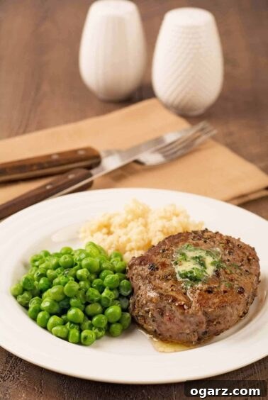 A mouth-watering peppercorn-crusted filet mignon, crowned with a pat of savory compound butter, is presented on a plate alongside creamy mashed potatoes and vibrant green peas. A folded napkin, fork, and knife are in the background.