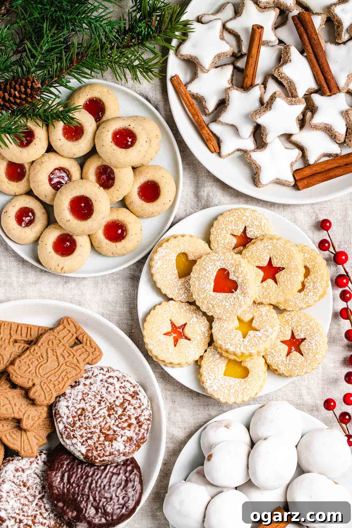 Starry Cinnamon Delights 6 A collection of German Christmas cookies on various plates garnished with pine branches and red berries. There are Cinnamon stars, garnished with cinnamon sticks, thumbprint cookies with strawberry jam, linzer cookies with strawberry jam and lemon curd, Gingerbread and spekulatius cookies and Pfeffernuesse.