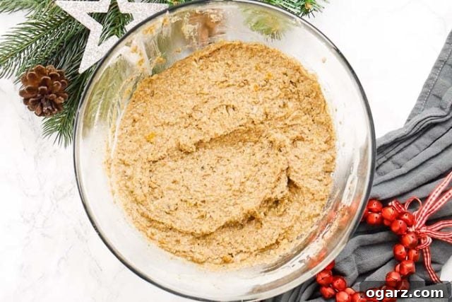 Authentic German Lebkuchen 4 A clear glass bowl filled with rich Lebkuchen dough, artfully placed next to a scattering of vibrant red jingle bells and fragrant pine branches, evoking a festive holiday atmosphere.