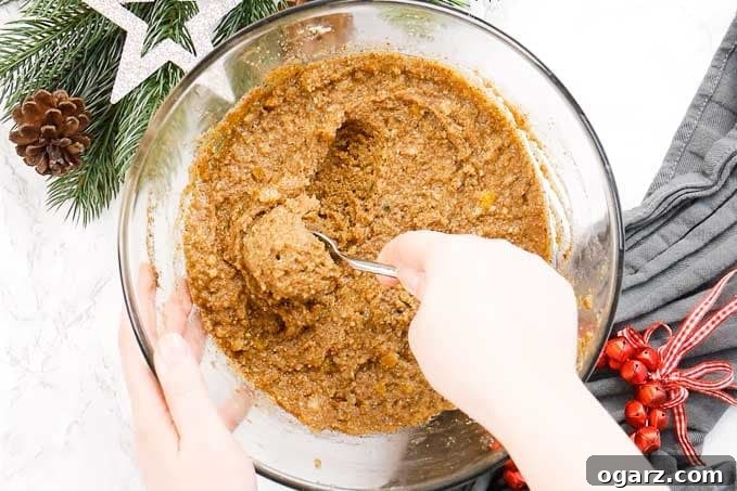 Authentic German Lebkuchen 5 A glass bowl of Lebkuchen dough sits amongst red jingle bells and pine branches, with a hand using a teaspoon to scoop out a portion of the thick batter.