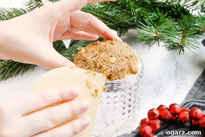 Authentic German Lebkuchen 6 A hand expertly shaping Lebkuchen dough on a wafer placed on an upside-down whiskey tumbler, using a dough card. Festive red jingle bells and pine branches create a cozy backdrop.