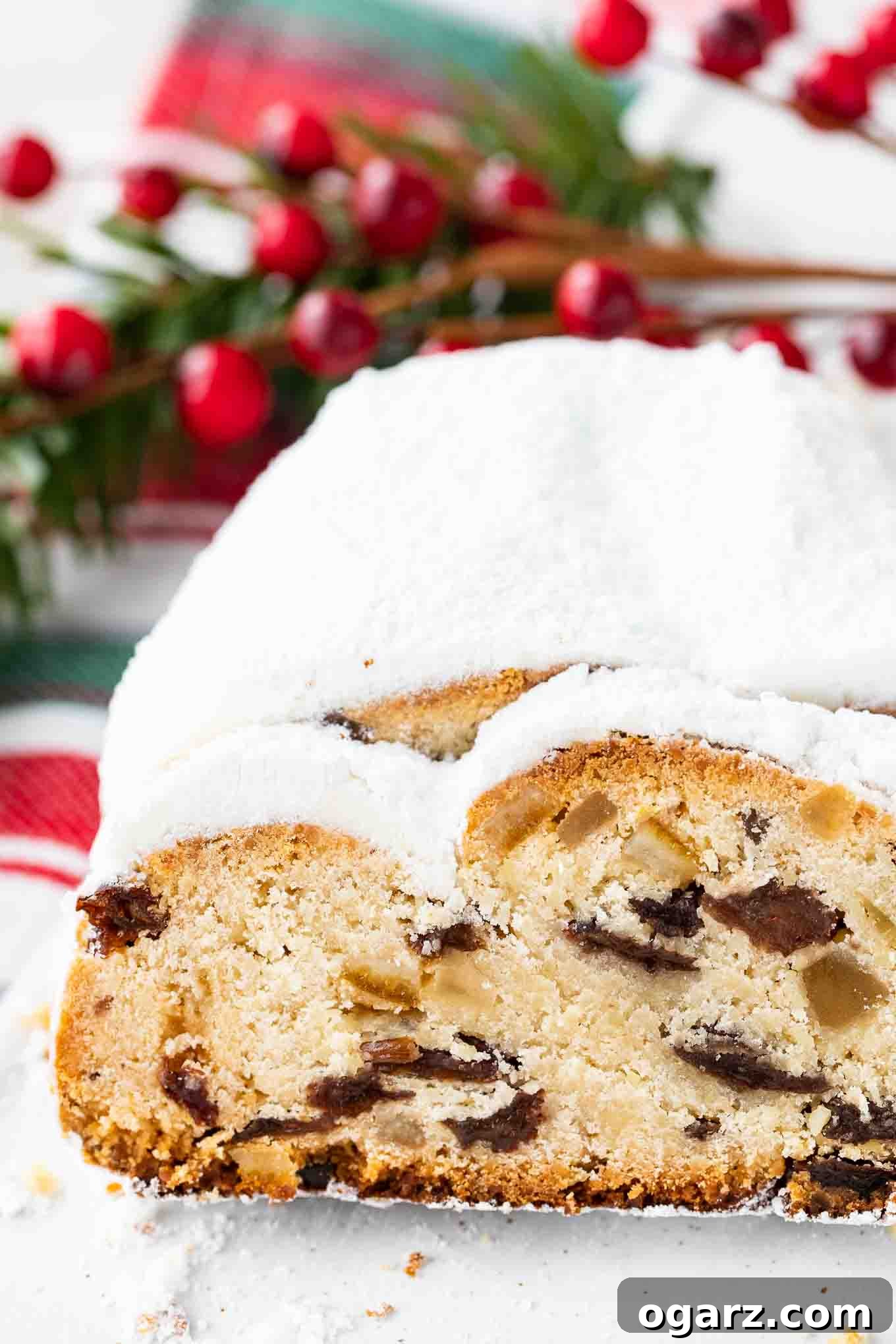 German Stollen is loaded with rum raisins, candied fruit, and nuts. This traditional German Christmas recipe is a very special treat that has a long history and is very popular during the Holidays. It needs a bit of time to develop its flavor but is totally worth the effort and tastes great with a cup of coffee! #GermanChristmasBaking #StollenRecipe Half a christstollen with a slice cut off on a red, white, and green dish towel, with fresh cranberries in the background.