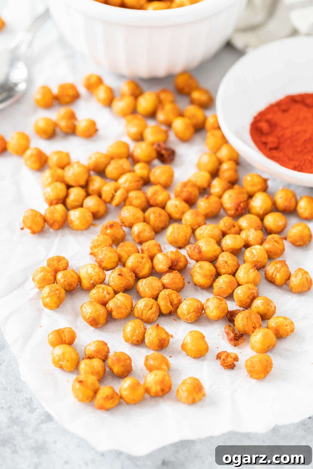 Crispy garbanzo beans on parchment paper next to a bowl of paprika, showcasing their golden-brown color and inviting texture.