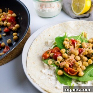 A white plate with spinach wrap with feta and chickpeas on it next to a pan with tomato chickpea mix. There's a jar with feta spread and half a lemon in the background.