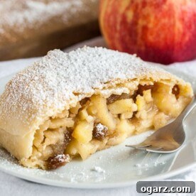A piece of apple strudel topped with powdered sugar on a white plate with a fork on a white dishcloth, with an apple in the background.