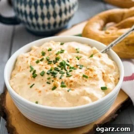A white bowl of Obatzda (German Beer Cheese Dip) with a spoon, garnished with chives on a wooden cutting board. There's a pretzel on a white dishtowel with a red stripe next to it and a beer stein in the background.