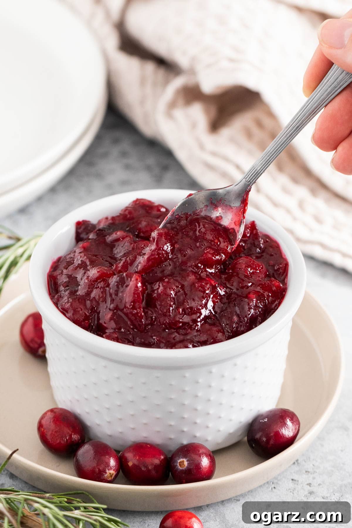 A woman's hand holding a spoon scooping out some cranberry sauce from a white bowl, showing its thick texture.