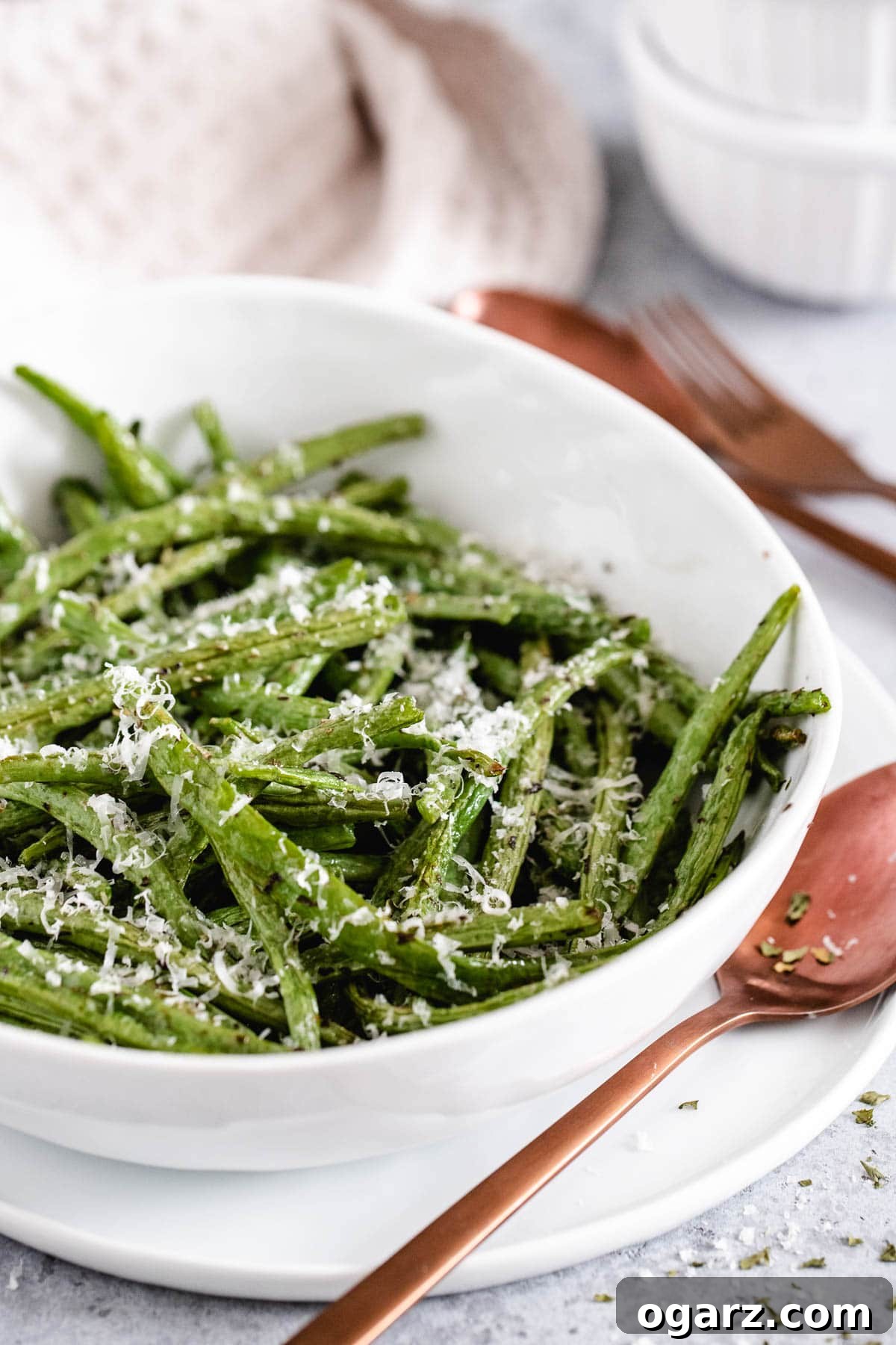 Crispy Air Fryer Green Beans 2 Cooked green beans in a white serving dish garnished with freshly grated Parmesan cheese, with a brown spoon resting beside the bowl on a light background.