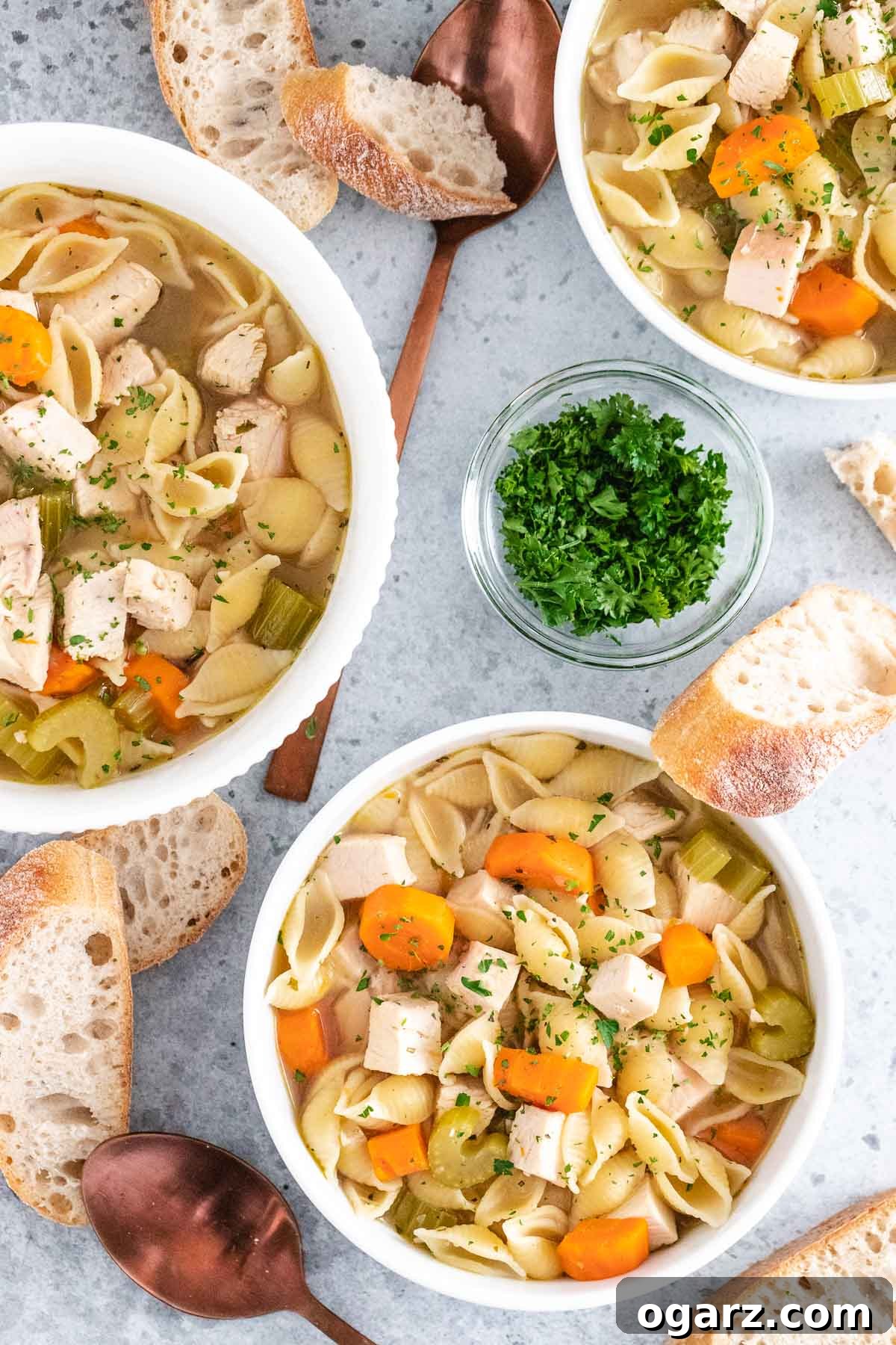 multiple servings of soup in white bowls on a grey background next to slices of bread, spoons, and a bowl of parsley