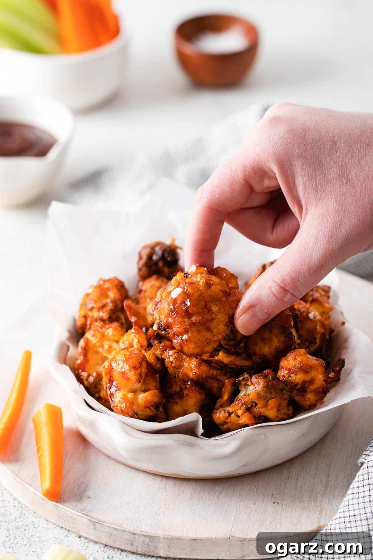 Crispy Air Fryer Cauliflower Bites 2 A female hand holding a crispy, sauce-coated Air Fryer Cauliflower Wing, with a bowl full of more wings in the background.