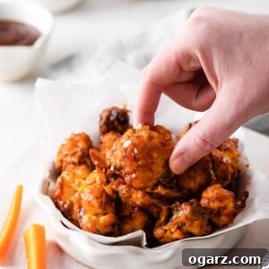 A female hand holding a cauliflower wing coated in BBQ sauce next to a white bowl filled with more wings
