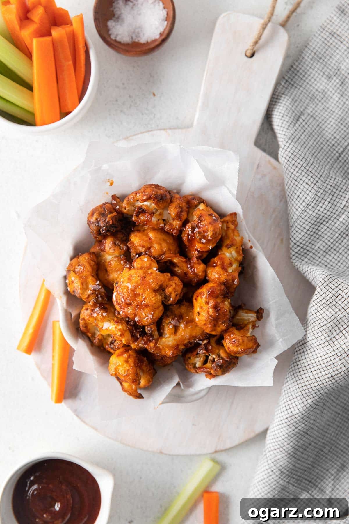 Crispy Air Fryer Cauliflower Bites 5 Cauliflower wings served in a stylish serving dish on a wooden board, accompanied by sliced vegetables and a small bowl of BBQ sauce.