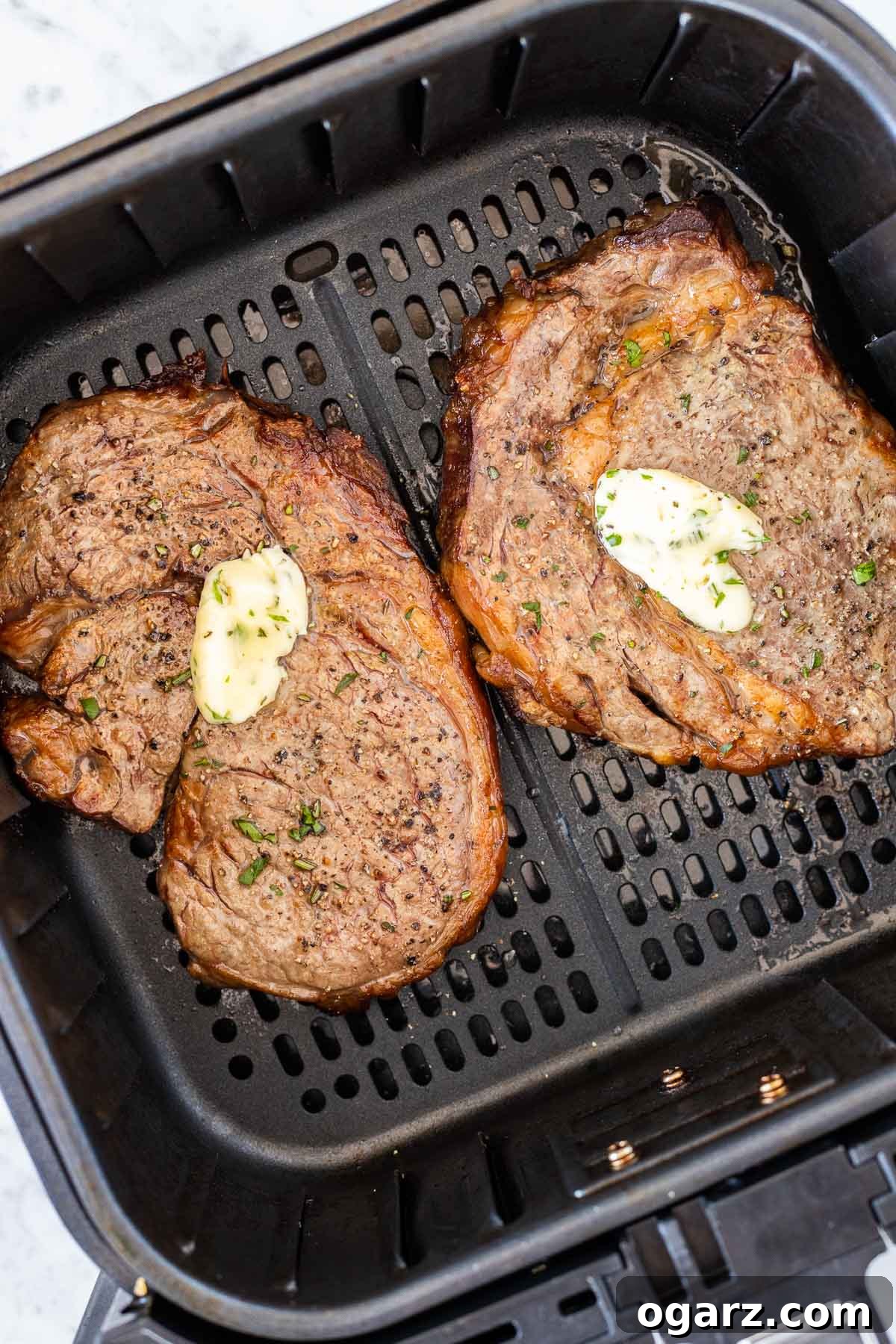 Two cooked ribeye steaks with a dollop of garlic butter on top, served in a black air fryer basket.