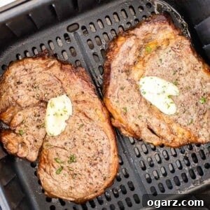 Two cooked steaks with a dollop of garlic butter on top in a black air fryer basket.