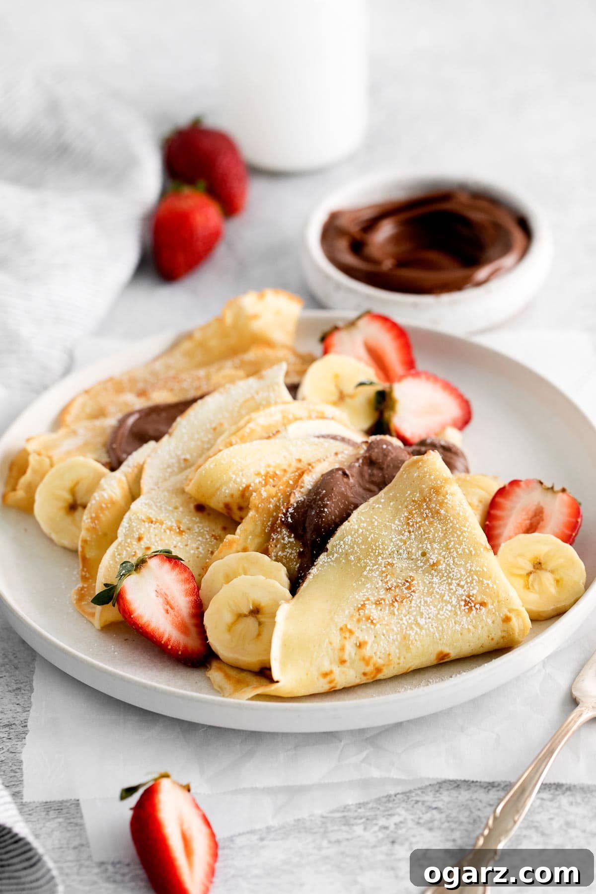 Effortless French Crepes 5 French crepes on a white plate garnished with fresh strawberries, banana slices, and a drizzle of Nutella