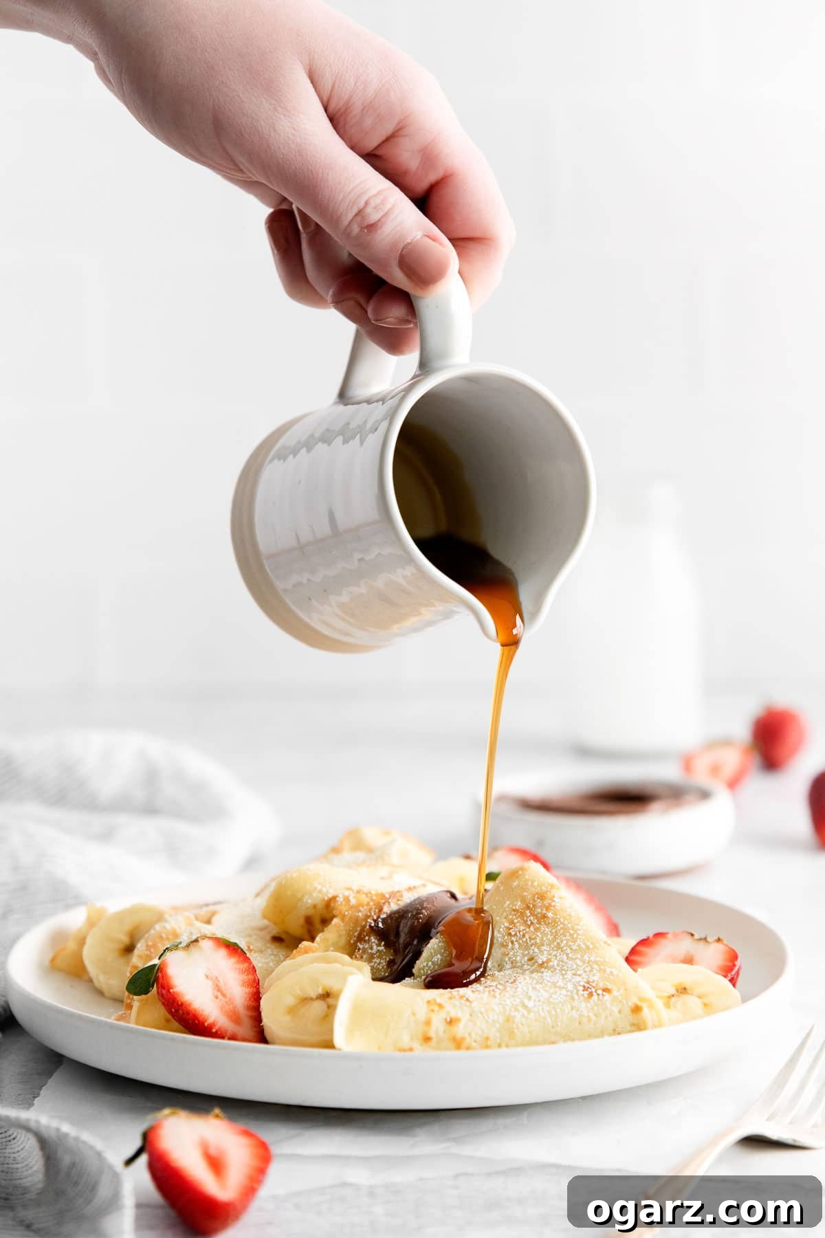 Effortless French Crepes 6 Maple syrup poured from a small jug over a stack of crepes on a white plate with fresh berries