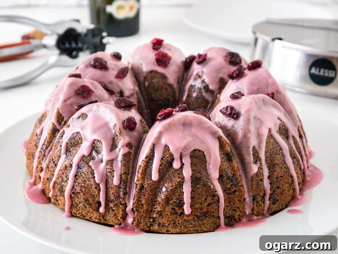 Chocolate Cranberry Bliss 6 A chocolate cranberry bundt cake with pink frosting, garnished with dried cranberries on a white plate. In the background, there's a stainless steel sugar bowl and a wine opener.