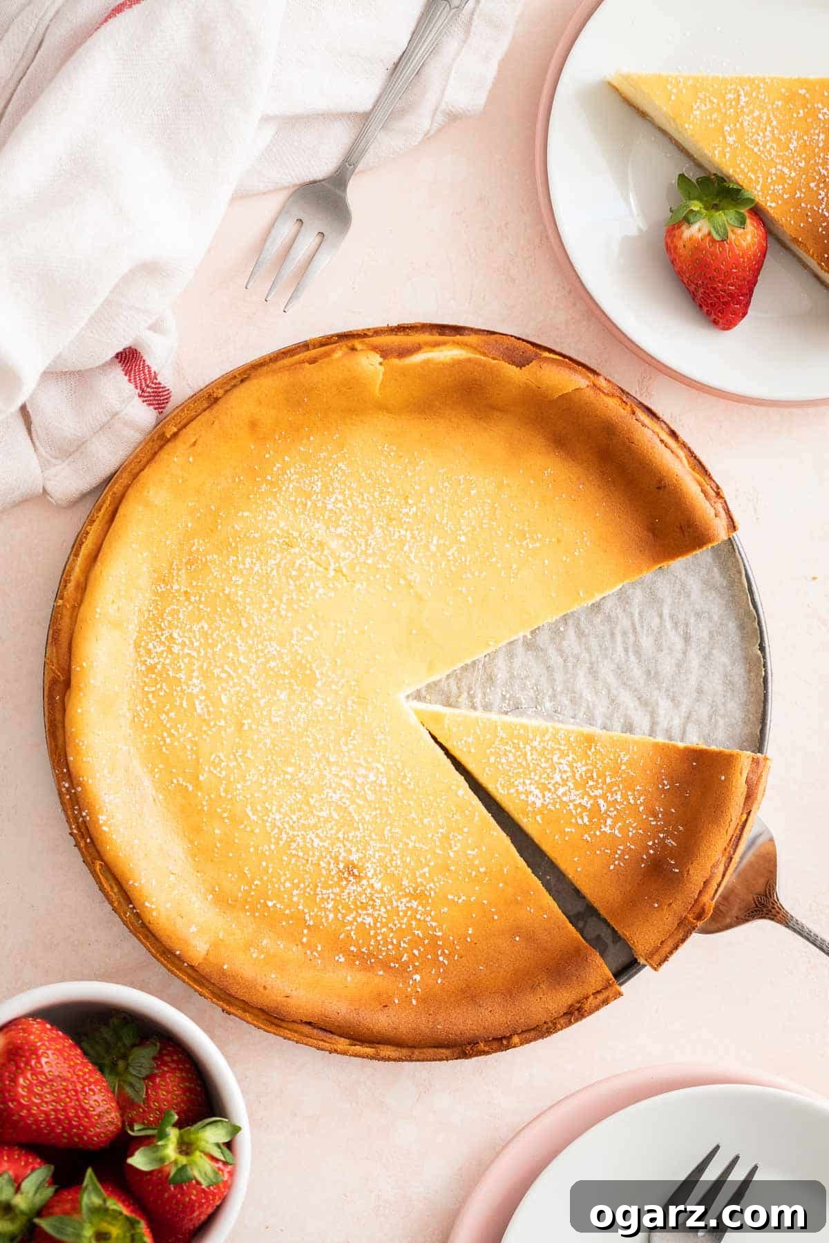 Käsekuchen: The German Cheesecake 6 A beautifully presented whole German Cheesecake resting on a pristine light pink surface, adorned with fresh strawberries and accompanied by elegant serving plates, ready to be enjoyed.