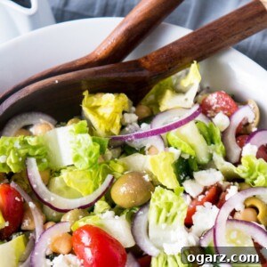 Close-up of a portion of Balsamic Chickpea Feta Greek Salad in a white bowl, showing the texture of chickpeas, olives, and red onions with a hint of balsamic dressing. Salad tongs are visible in the background.