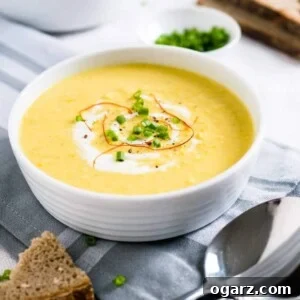 A white bowl of curried cream of corn soup, garnished with sour cream, chili threads and chives on a grey dishtowel. There's a piece of bread in front of it and a spoon next to it. Another bowl of soup, a small white bowl of chives and more bread is in the background.