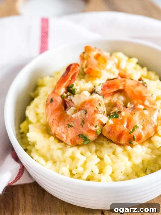 Saffron Shrimp Risotto is really easy to make, super creamy, and ready in only 30 minutes! A variation of the famous Risotto Milanese made with garlic shrimp that is quick enough to prepare for weeknights but special enough for date night. Close-up of a white bowl with saffron risotto with fried shrimp on a white dish towel with a red stripe. The dish is beautifully garnished, highlighting the golden hue of the risotto and the perfectly cooked pink shrimp.