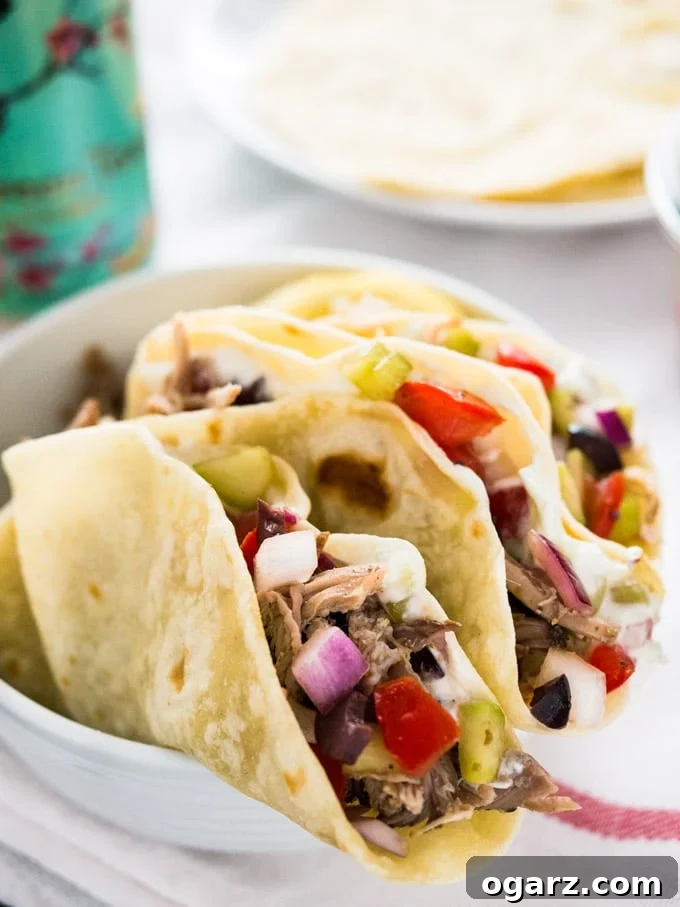 A white bowl greek style tacos, filled with greek style pulled pork, salad and tzatziki. There's a white plate of tortillas and a can of Arizona ice tea in the background.