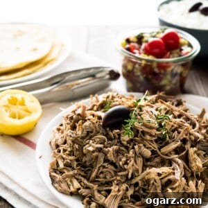 Mediterranean Slow Cooker Pulled Pork Gyros 7 A plate of pulled pork, garnished with an olive and thyme sprigs on a white dishtowel with a red stripe. There's a jar of greek salad, a bowl of tzatziki, half a lime, some tongs and a plate of tortillas in the background.