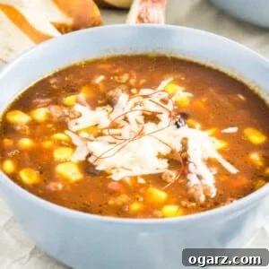 A grey bowl of chili con carne with beer and corn topped with chili threads and grated cheese. There are slices of pretzel buns in the background.