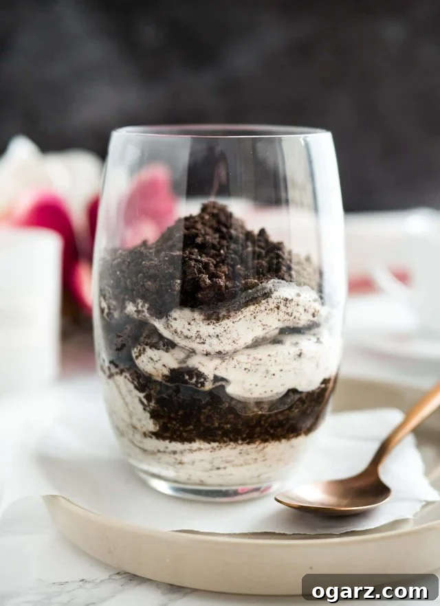A close-up view of a beautifully layered No Bake Oreo Cheesecake Parfait, topped with generous Oreo crumbs, presented in a clear glass on parchment paper, resting on a gray plate with a bronze spoon nearby.