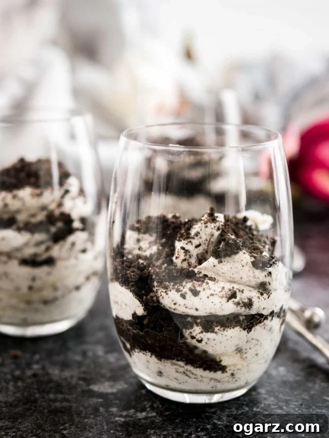 Two elegant glasses filled with No Bake Oreo Cheesecake Parfaits, featuring distinct layers of creamy filling and crushed cookies, garnished with extra Oreo crumbs. They are set on parchment paper atop a gray plate, accompanied by a stylish bronze spoon.