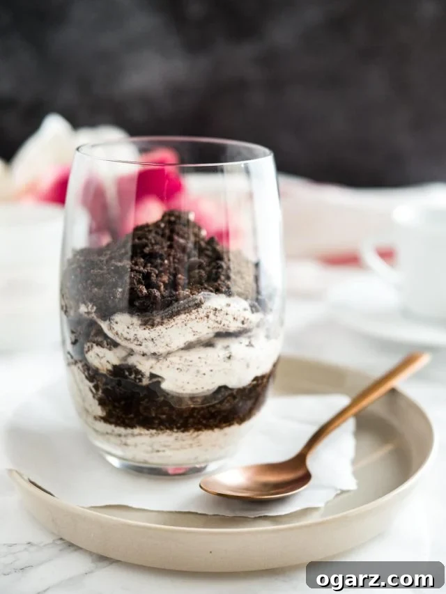 A single, enticing glass of No Bake Oreo Cheesecake Parfait, showcasing its creamy layers and crunchy Oreo topping. The glass sits on parchment paper on a gray plate, with a bronze spoon invitingly placed beside it.
