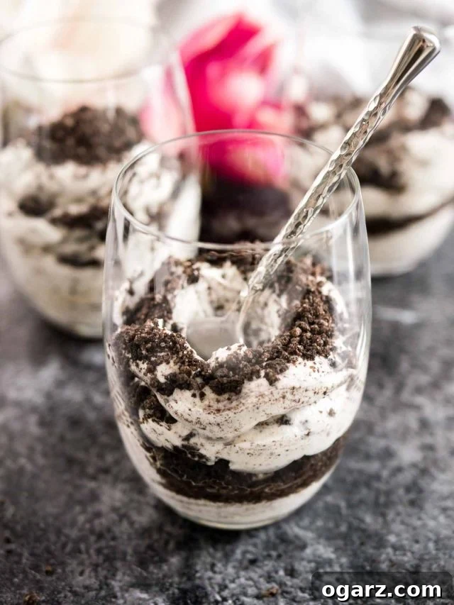 A pair of beautifully arranged No Bake Oreo Cheesecake Parfaits, showcasing their inviting layers and crumbly toppings. They are artfully placed on parchment paper, on a sleek gray plate, with a bronze spoon poised for enjoyment.