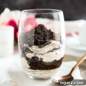 Close-up of a glass of oreo cheesecake parfaits topped with oreo crumbs on a piece of parchment on a grey plate with a bronze spoon next to it.