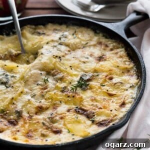 A cast-iron pan with gorgonzola potatoes au gratin wit a spoon digging into it on a wooden table. There's a white dishtowel next to it and a stack of plates with forks in the background.