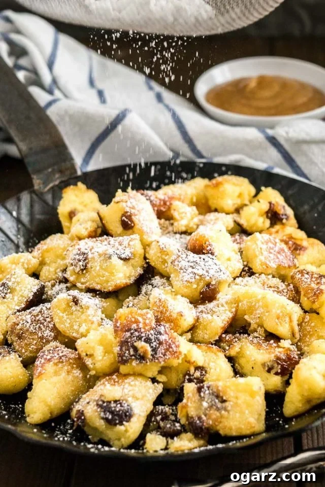 Kaiserschmarrn (Scrambled Pancakes) is an Austrian dessert that's also very popular in Bavaria! A sweet fluffy pancake made with rum-soaked raisins is torn into bite-sized pieces, caramelized, and served sprinkled with powdered sugar, applesauce, and preserves. This easy dessert recipe also tastes great for brunch and is loved by kids and adults alike! #Pancakes #DessertRecipes #Kaiserschmarrn #GermanRecipes #ComfortFood A pan with Kaiserschmarrn with powdered sugar on top on a white and blue dish towel with a white bowl of apple sauce in the background