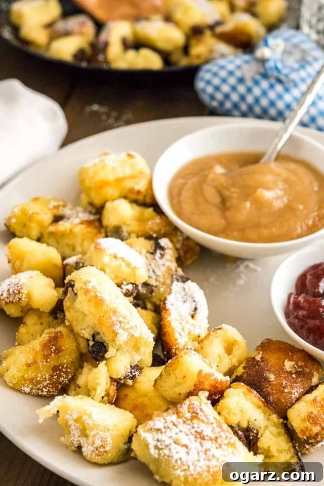 Kaiserschmarrn (Scrambled Pancakes) is an Austrian dessert that's also very popular in Bavaria! A sweet fluffy pancake made with rum-soaked raisins is torn into bite-sized pieces, caramelized, and served sprinkled with powdered sugar, applesauce, and preserves. This easy dessert recipe also tastes great for brunch and is loved by kids and adults alike! #Pancakes #DessertRecipes #Kaiserschmarrn #GermanRecipes #ComfortFood A plate with Kaiserschmarrn topped with powdered sugar with two small white bowls, one with apple sauce, one with plum puree on a wooden table.