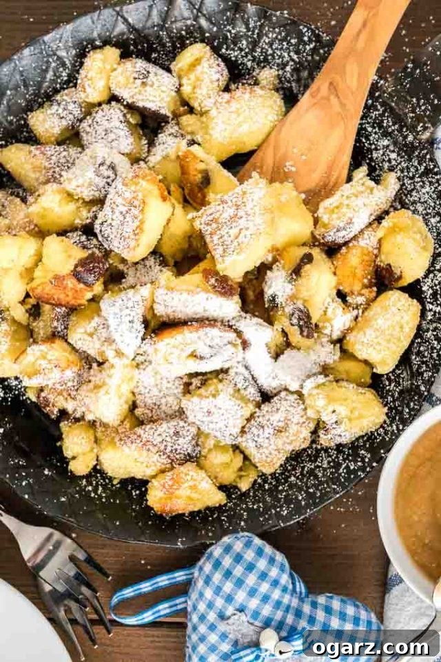 Kaiserschmarrn (Scrambled Pancakes) is an Austrian dessert that's also very popular in Bavaria! A sweet fluffy pancake made with rum-soaked raisins is torn into bite-sized pieces, caramelized, and served sprinkled with powdered sugar, applesauce, and preserves. This easy dessert recipe also tastes great for brunch and is loved by kids and adults alike! #Pancakes #DessertRecipes #Kaiserschmarrn #GermanRecipes #ComfortFood A pan of Kaiserschmarrn with raisins in a pan, topped with powdered sugar, with a wooden spatula in it. There\'s a fork, a white and blue plaid towel and a small white bowl of apple sauce next to it.