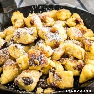 The Emperor's Shredded Pancake 2 Close-up of a pan with Kaiserschmarrn with powdered sugar on top on a white and blue dish towel with a white bowl of apple sauce in the background