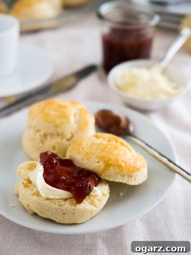 Effortless English Scones 2 A pristine white plate showcases freshly baked, golden English scones, accompanied by a delicate spoon of shimmering marmalade. One scone is elegantly halved, revealing layers generously spread with velvety clotted cream and vibrant strawberry jam. In the soft-focus background, a small white bowl brimming with clotted cream and a rustic jar of strawberry jam complete this inviting British afternoon tea scene.