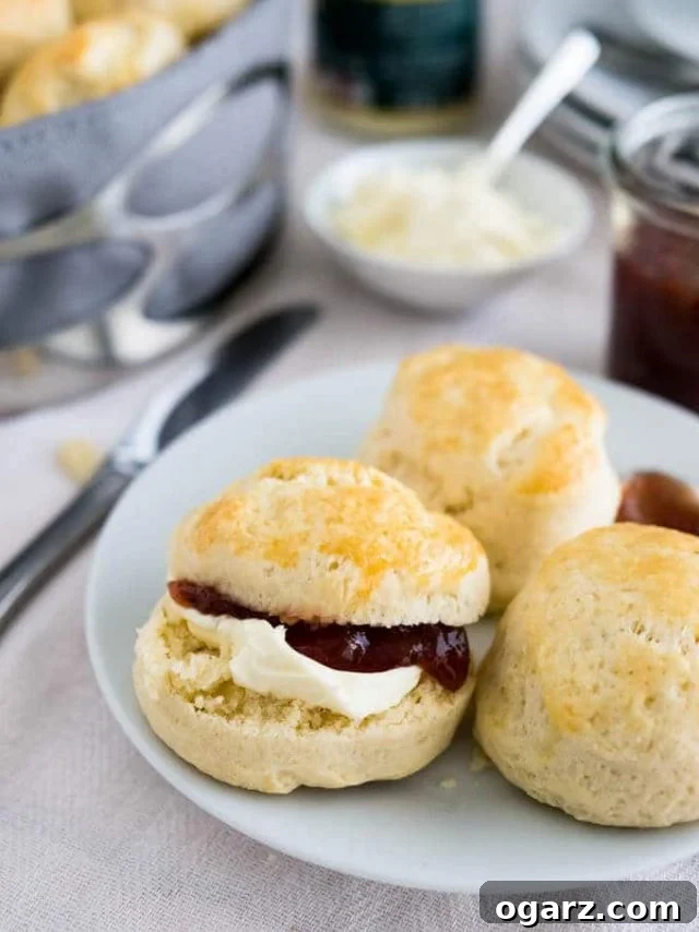 Effortless English Scones 3 A beautifully arranged white plate showcasing a selection of freshly baked English scones, ready for serving. A small spoon of marmalade rests elegantly on the side. One of the golden-brown scones is artfully sliced in half, revealing its tender interior, and is lavishly layered with thick, luxurious clotted cream and glistening strawberry jam. In the background, a pristine white bowl is filled with extra clotted cream, alongside a charming glass jar of strawberry jam, emphasizing the classic British pairing.