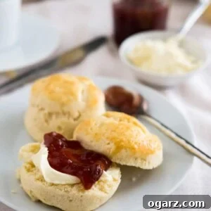 Effortless English Scones 7 A white plate with perfectly baked English scones, a spoonful of marmalade, and one scone halved with clotted cream and strawberry jam. A small bowl of clotted cream and jam jar are in the background.