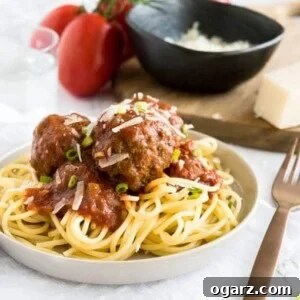 A grey plate with spaghetti and Italian Meatballs with tomato sauce on a white dishtowel. There's a bronze fork next to it and a glass of water, some tomatoes on the vine and a wooden cutting board with a black bowl of grate Parmiggiano in the background.